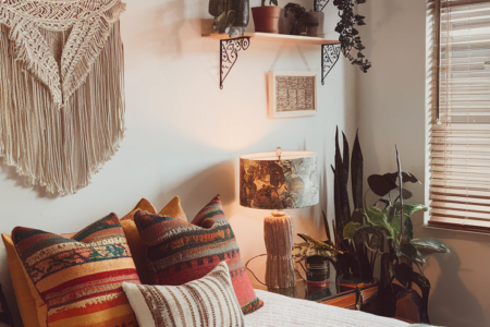 A photo of a cozy bedroom with a deep blue wall. The bed has blue bedding and patterned pillows, and is adorned with a multicolored blanket. To the left, a window lets in natural light and reveals greenery outside. Above the bed hangs a unique woven lantern. The wall behind the bed is decorated with multiple framed pictures, including a sunset scene and various artworks. On the right, a wooden cabinet holds a hat and some decorative items. A small table beside the bed holds a candle and a few books.