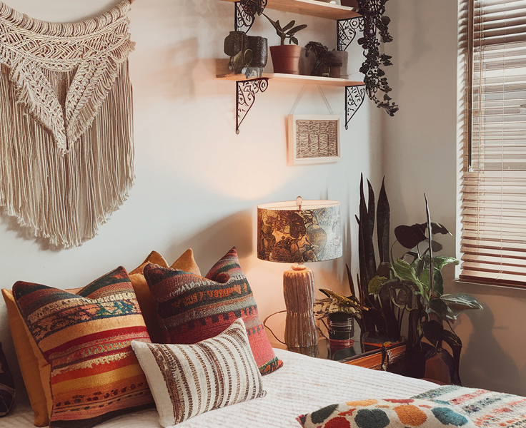 A photo of a cozy bedroom with a deep blue wall. The bed has blue bedding and patterned pillows, and is adorned with a multicolored blanket. To the left, a window lets in natural light and reveals greenery outside. Above the bed hangs a unique woven lantern. The wall behind the bed is decorated with multiple framed pictures, including a sunset scene and various artworks. On the right, a wooden cabinet holds a hat and some decorative items. A small table beside the bed holds a candle and a few books.