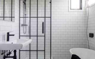 A modern bathroom with white tiles. On the left, there's a wall-mounted sink with a black faucet. Adjacent to it is a glass-enclosed shower area with a black frame. Above the sink, there's a rectangular mirror. On the right, there's a freestanding bathtub with a white finish. The floor is also covered in white tiles, and there's a black towel placed near the bathtub. A window with a curtain is visible on the top right, allowing natural light to illuminate the space.