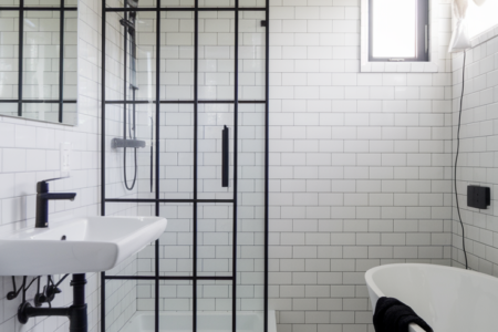 A modern bathroom with white tiles. On the left, there's a wall-mounted sink with a black faucet. Adjacent to it is a glass-enclosed shower area with a black frame. Above the sink, there's a rectangular mirror. On the right, there's a freestanding bathtub with a white finish. The floor is also covered in white tiles, and there's a black towel placed near the bathtub. A window with a curtain is visible on the top right, allowing natural light to illuminate the space.